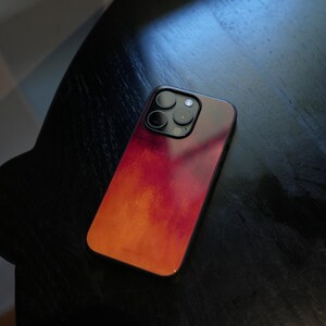 Op de afbeelding: Een smartphonehoesje met een gradiëntontwerp in tinten rood en oranje. De hoes heeft een zwarte rand en een cameramodule met drie lenzen. De hoes ligt op een donkere ondergrond.