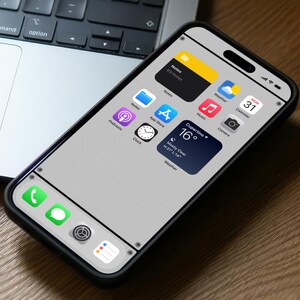 May include: A black smartphone with a gray screen displaying various app icons, including Notes, Weather, Calendar, Music, Camera, Podcasts, Clock, and App Store. The phone is on a wooden surface with a laptop and white earbuds in the background. The phone screen also shows the current weather in Cupertino, California, with a temperature of 16 degrees Celsius and a mostly clear sky.
