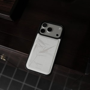 Op de afbeelding: Een smartphonehoesje met een zwart frame en een wit achterpaneel met een subtiel, lichtgrijs bamboebladpatroon. Het hoesje bevat een verhoogde uitsparing voor de camera met drie ronde lensopeningen en een kleine flitsopening rechtsboven.