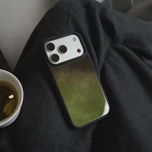 May include: A black phone case with a green and brown watercolor design rests on dark gray fabric. The phone case has a black border and a camera module with multiple lenses. A white teacup with tea is visible.