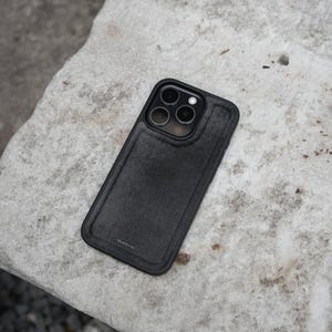May include: A black smartphone case with a textured surface. The case has a rectangular shape with a camera cutout and a raised border around the edges. The case is resting on a light gray, textured surface.