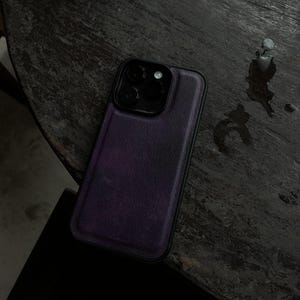 May include: A purple leather phone case with a black camera module. The case is on a dark wooden surface. The phone case has a textured appearance and a black border.