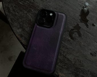 Può includere: Una custodia per telefono in pelle viola con un modulo fotocamera nero. La custodia è su una superficie di legno scuro. La custodia ha un aspetto strutturato e un bordo nero.
