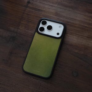 May include: A green and black phone case with a textured finish. The case has a black border and a green center. The phone's camera lenses are visible at the top. The case is on a dark wooden surface.