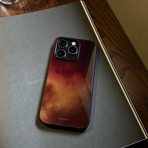 以下が含まれることがあります： ブルゴーニュからゴールドにグラデーションするデザインのスマートフォンケース。ケースには黒い縁とカメラの切り抜きがあります。「bluestone」の文字がケースにプリントされています。ケースは灰色の表面にあります。