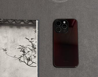 Può includere: Uno smartphone bordeaux con una fotocamera a tripla lente è posizionato su una superficie grigia. La finitura lucida del telefono riflette la luce. A sinistra, una stampa incorniciata in bianco e nero di una pianta.