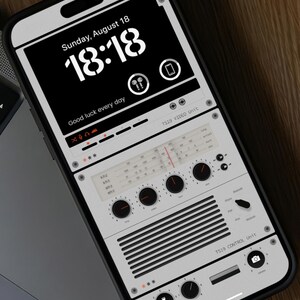 May include: A white smartphone with a black screen displaying a digital clock with the time 18:18 and the date Sunday, August 18. The text "Good luck every day" is displayed below the clock. The screen also shows a stylized image of a vintage radio with knobs and dials.