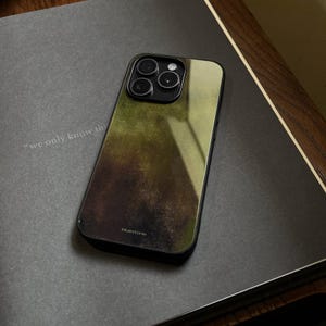 May include: A smartphone case with a gradient design in shades of green and brown. The case has a black border and a camera cutout. The word "bluestone" is printed at the bottom. The phone case is resting on a gray surface.