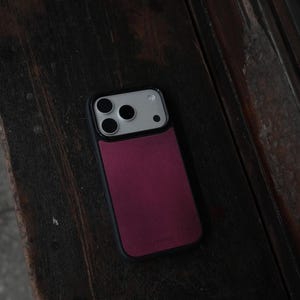 May include: A smartphone case with a black frame and a burgundy back panel. The case has a camera cutout with three lenses and a flash. The phone case is resting on a dark wooden surface.