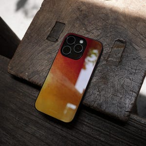 May include: A smartphone case with a vibrant abstract design in shades of red, orange, and yellow. The case has a black border and features a camera module with multiple lenses. The case is resting on a weathered wooden surface.