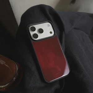 May include: A smartphone case with a burgundy-colored back and a black frame. The case has a glossy finish and a camera module with three lenses. The phone case is resting on a dark fabric.