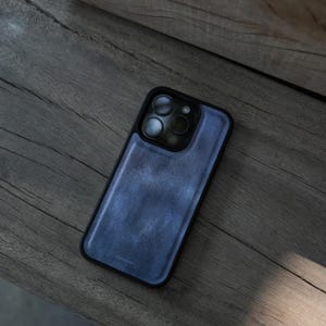 May include: A blue leather phone case with a black border. The case is on a wooden surface. The phone case has a textured appearance and covers the back and sides of the phone, with cutouts for the camera lenses.