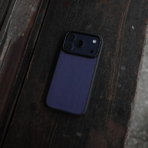 May include: A dark blue smartphone case with a black camera module. The case has a textured finish and is set against a dark wooden surface. The phone case is designed to protect the phone.