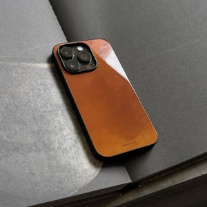 May include: A brown and black smartphone case with a glossy finish. The case has a rectangular shape with rounded corners and a camera cutout. The case is resting on a dark gray surface.
