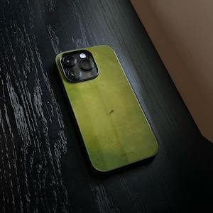 Op de afbeelding: Een smartphone met een beschermhoes in een gladde, olijfgroene afwerking die subtiele, gevlekte textuurvariaties vertoont. Het apparaat heeft een opvallende triple-lens camera-array in de linkerbovenhoek, omrand door een zwart frame.