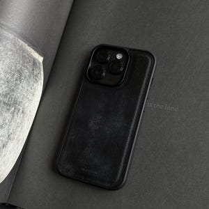 May include: A black leather phone case with a camera lens protector. The case has a textured surface and is designed to fit a smartphone. The phone case is on a dark gray surface.