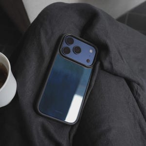 May include: A smartphone in a black case with a blue gradient back. The phone has three camera lenses and a flash. The phone is resting on a dark gray fabric surface.