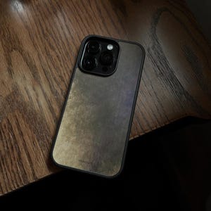 May include: A smartphone case with a textured, metallic finish in shades of gray and gold. The case has a black border and a camera module with three lenses and a flash. The brand name is printed at the bottom.