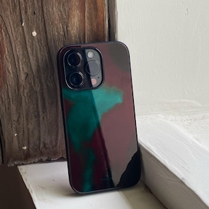 May include: A smartphone case with a dark, glossy finish. The case features a camouflage-like pattern in shades of burgundy, teal, and black. The case has a camera cutout with three lenses and a flash.