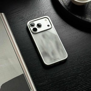 Puede incluir: Una funda rectangular para smartphone con marco metálico plateado y panel trasero semitransparente esmerilado con un sutil patrón de mármol gris. El módulo de cámara es plateado con tres aberturas circulares negras para lentes.