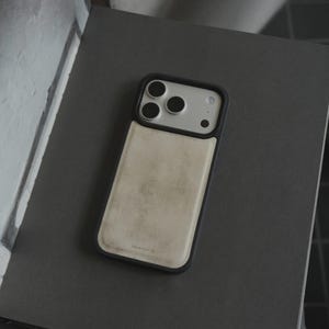 May include: A smartphone case with a black frame and a light beige leather back. The case has a camera cutout with three lenses. The phone case is resting on a dark gray surface.