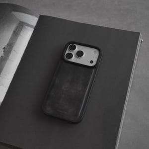 May include: A black smartphone case with a textured finish, featuring a camera module with three lenses. The case is resting on a dark gray open book, creating a minimalist aesthetic. The image is in grayscale.