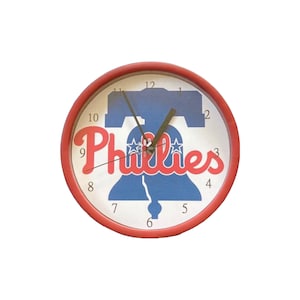 May include: A red and white wall clock with a Philadelphia Phillies logo. The logo features a blue Liberty Bell with two stars and the word "Phillies" in red.