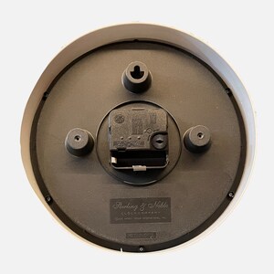 K&ouml;nnte beinhalten: Die R&uuml;ckseite einer wei&szlig;en runden Uhr mit schwarzem Zifferblatt. Die Uhr hat ein schwarzes Uhrwerk und ein Etikett mit der Aufschrift "Sterling & Noble Clock Company" und "Made in China".