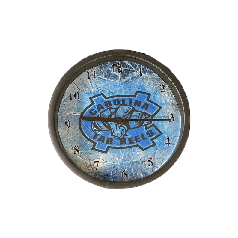 K&ouml;nnte beinhalten: Eine schwarz-wei&szlig;e Uhr mit einem blau-wei&szlig;en, rissigen Eisdesign. Das Zifferblatt der Uhr zeigt das Logo der University of North Carolina Tar Heels mit dem Text "Carolina Tar Heels".