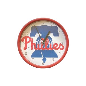 May include: A round wall clock with a red frame. The clock face features the Philadelphia Phillies logo, including a blue Liberty Bell and the word "Phillies" in red script. Black clock hands indicate the time.