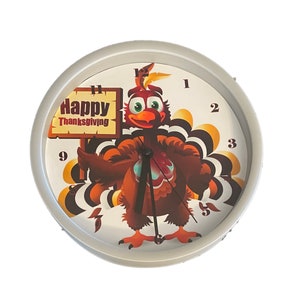 May include: A round, silver-colored clock with a cartoon turkey holding a sign that says "Happy Thanksgiving". The turkey is brown and orange with a red wattle and a blue and white patterned tail.