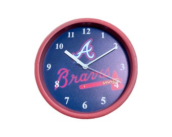 Puede incluir: Reloj de pared redondo con marco rojo y esfera azul marino. El reloj presenta el logotipo de los Atlanta Braves, con el nombre del equipo y un diseño de bate de béisbol. Las manecillas del reloj son blancas.