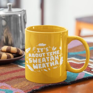 May include: A yellow ceramic mug with a white design that says "It's About Time Sweatah Weatha" with leaf illustrations. The mug is on a table with a brown and black patterned placemat.