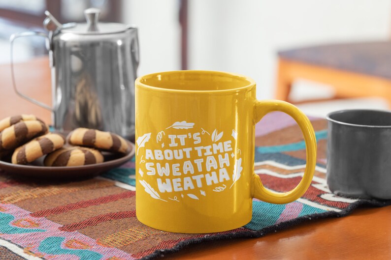 May include: A yellow ceramic mug with a white design that says "It's About Time Sweatah Weatha" with leaf illustrations. The mug is on a table with a brown and black patterned placemat.