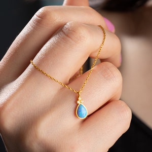 Puede incluir: Un delicado collar de cadena de oro con un colgante en forma de l&aacute;grima que presenta una piedra de &oacute;palo azul y peque&ntilde;as piedras preciosas transparentes.