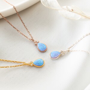 Puede incluir: Tres delicados collares con colgantes de &oacute;palo en forma de l&aacute;grima y peque&ntilde;os detalles de diamantes. Los collares est&aacute;n sobre una superficie blanca con una ramita de flores secas.