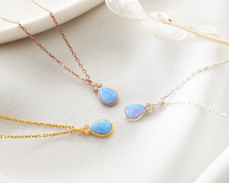 Puede incluir: Tres delicados collares con colgantes de &oacute;palo en forma de l&aacute;grima y peque&ntilde;os detalles de diamantes. Los collares est&aacute;n sobre una superficie blanca con una ramita de flores secas.