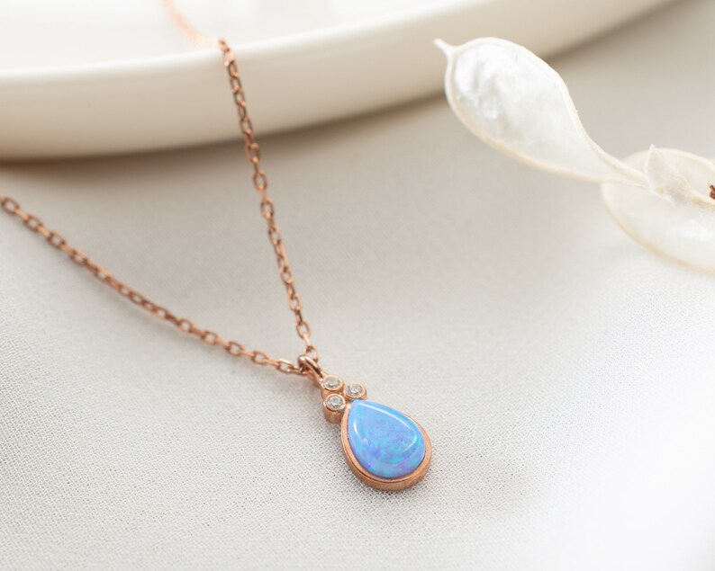 Puede incluir: Un delicado collar de oro rosa con un colgante de opalo azul en forma de l&aacute;grima adornado con peque&ntilde;as piedras transparentes.