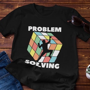 Può includere: Maglietta nera con un design a cubo di Rubik colorato e il testo "PROBLEM SOLVING".