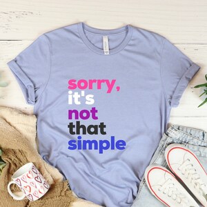 Puede incluir: Una camiseta azul claro con el texto "sorry, it's not that simple" en letras rosa, azul y negras.