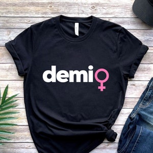 May include: Black t-shirt with the word "demi" in white and a pink female symbol. The shirt is displayed on a wooden surface with a pair of blue jeans, a black cap, and a green plant.