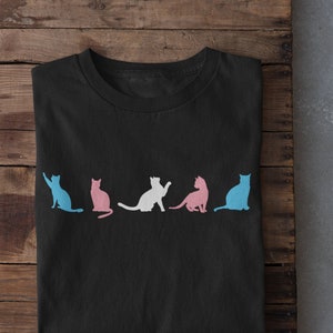 May include: A black t-shirt featuring a line of five cat silhouettes in various colours. The cats are arranged in a row, facing the right side of the image. The cats are blue, pink, white, pink, and blue.