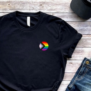 Può includere: Maglietta nera con un simbolo di freccia bianca e arcobaleno in un cerchio sul petto sinistro.