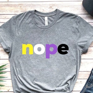 May include: Gray t-shirt with the word "nope" printed in yellow, purple, and black letters. The shirt is laid out on a wooden surface with a pair of jeans, a hat, and a plant.