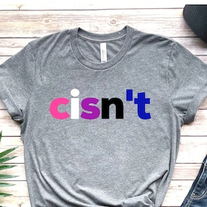 May include: A grey t-shirt with the text "cisn't" printed in pink, white, purple, and blue letters. The shirt is on a wooden surface with a green plant, a pair of blue jeans, and a black baseball cap.
