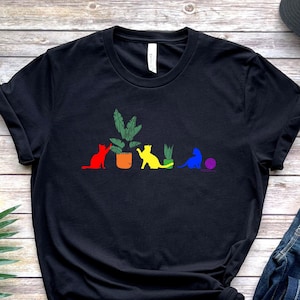 May include: Black t-shirt with a graphic of five cats in a row. The cats are sitting in front of potted plants. The cats are colored in a rainbow pattern. The shirt is on a wooden surface with a pair of jeans and a black baseball cap.