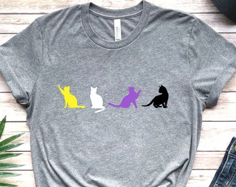 Op de afbeelding: Een grijs T-shirt met een ontwerp van drie katten op een rij. De katten zijn geel, paars en zwart. Het shirt ligt op een houten oppervlak met een plant, jeans en een pet.