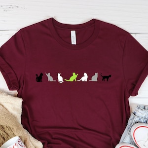 May include: Maroon t-shirt featuring a row of stylized cat silhouettes in black, gray, white, and lime green. The shirt is displayed with a beige scarf, a mug with heart designs, and white sneakers.