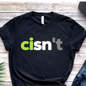 Puede incluir: Camiseta negra con el texto "cisn't" en verde, blanco y gris. La camiseta se combina con vaqueros azules, una gorra negra y zapatillas blancas. Presentado sobre una superficie de madera.