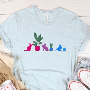 May include: A light blue t-shirt with a graphic of five cats in a row, each with a potted plant. The cats are in different colors, and the plants are in different shades of green. The text "HEATHER ICE BLUE" is at the bottom of the image.